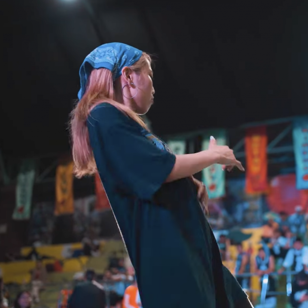 Nono Sasai, a young woman with long hair, wearing a blue headband and black oversized shirt, performs a dance move with her hand raised and gesturing to the side. The background shows an audience watching from bleachers adorned with colorful banners and a wall with vibrant murals. The setting is an indoor venue with a lively, engaging atmosphere.